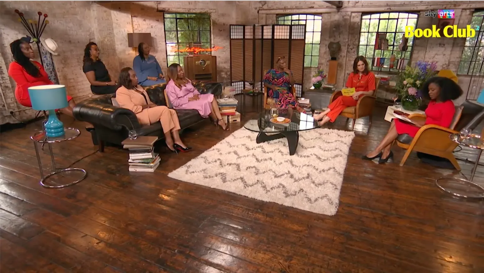 A woman in a red dress reads a book to a group of women sat on sofas and chairs arranged in a semi-circle